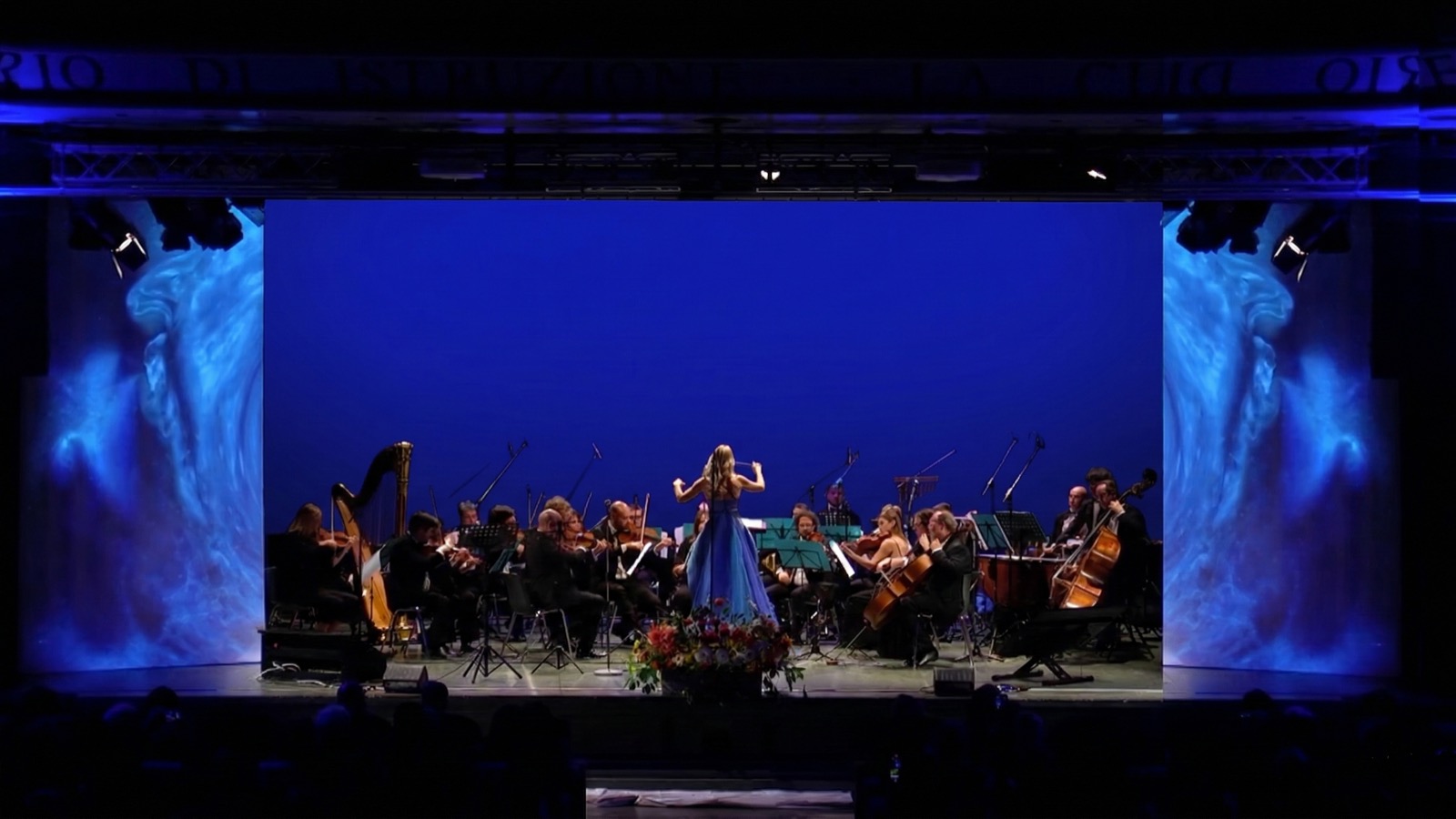 Orchestra Ars Armonica con tempesta d'acqua proiettata sulle quinte e fondale blu intenso — scenografia digitale per il Concerto per la Natura, Sala Oggioni Bergamo 2022, SUONO E LUCE