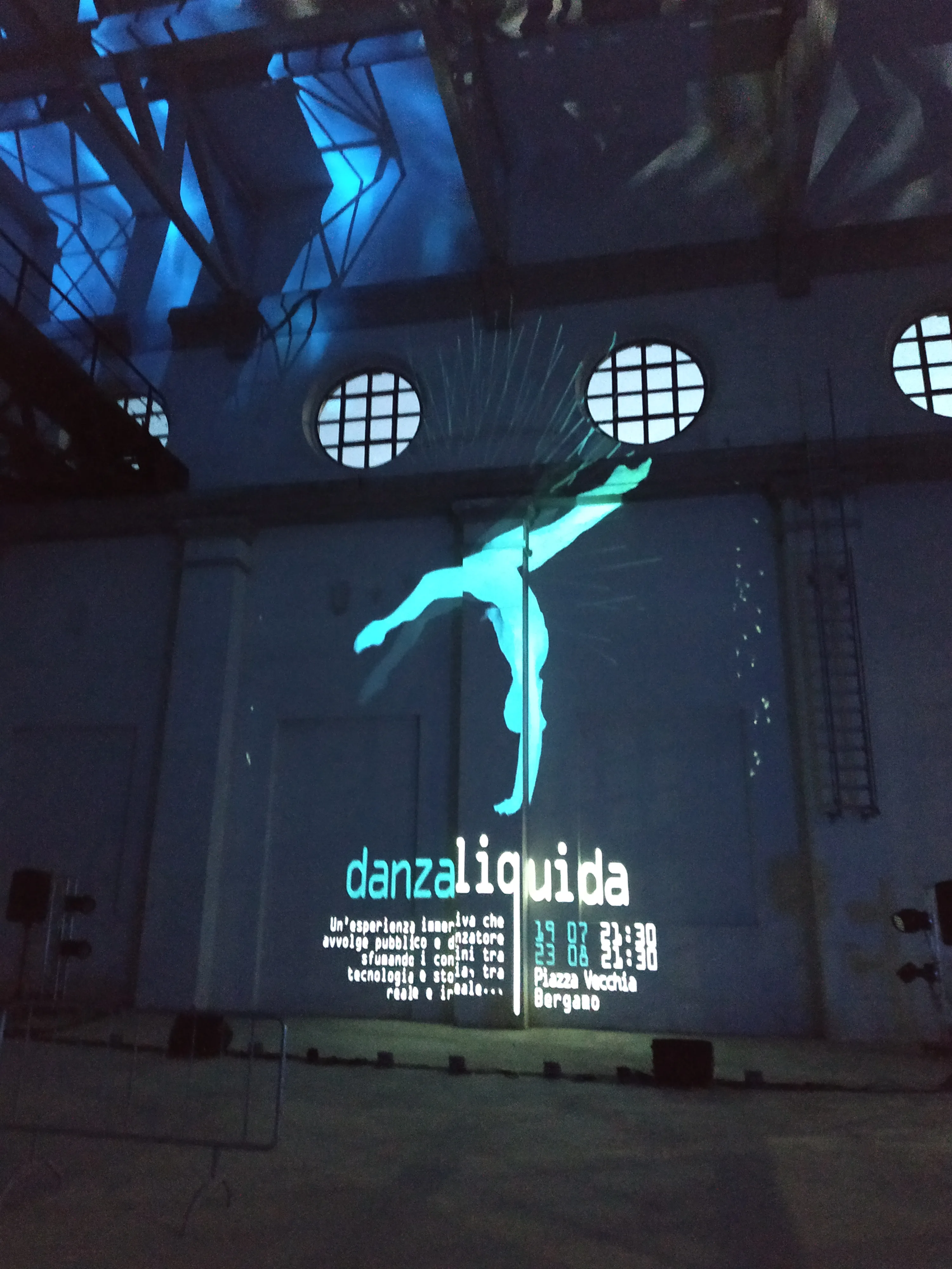 Maxiproiezione della locandina Danza Liquida sulla parete della Ex Centrale ENEL di Daste e Spalenga — silhouette ciano della danzatrice, oblò industriali e strutture in acciaio, SUONO E LUCE