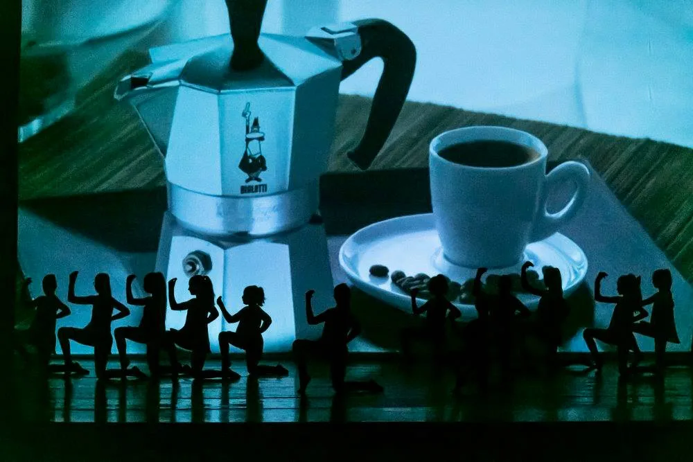 Silhouette di danzatrici contro moka Bialetti e tazzina di caffè proiettate sul fondale — videoproiezione scenica SUONO E LUCE