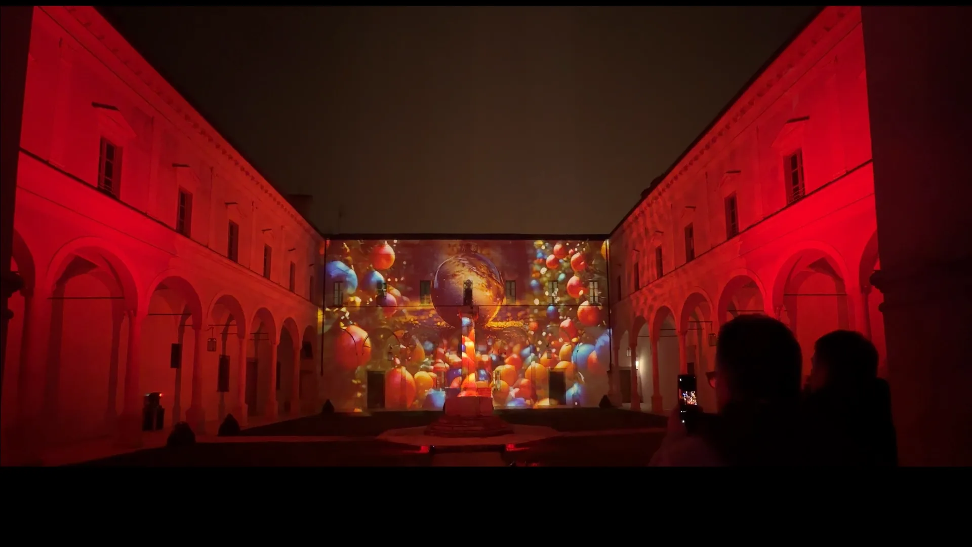 Proiezioni rosse con sfere luminose e pubblico nel chiostro dell'Abbazia — auguri di Natale, serata di solidarietà 19 dicembre 2025, videomapping SUONO E LUCE