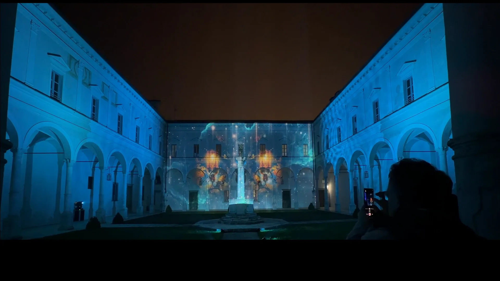 Chiostro dell'Abbazia in blu con lanterne arancio e stelle proiettate sulla parete frontale — serata di solidarietà 19 dicembre 2025, videomapping SUONO E LUCE